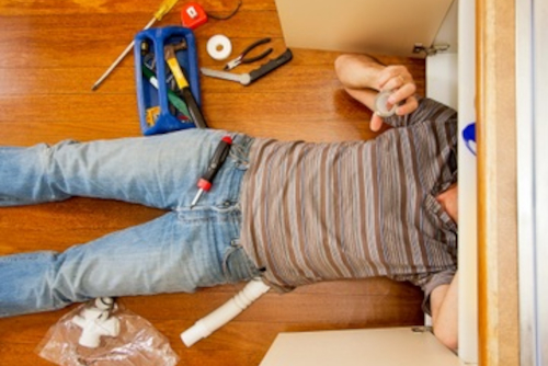 man-doing-sink-plumbing-repair-under-kitchen-cabinet Man lying under kitchen sink with tools, repairing pipes and drain connection